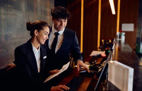 Happy hotel managers through reservation list on desktop PC at reception desk.