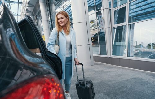 Smiling pleased woman with trolley suitcase closing door of taxicab parked before airport terminal building
